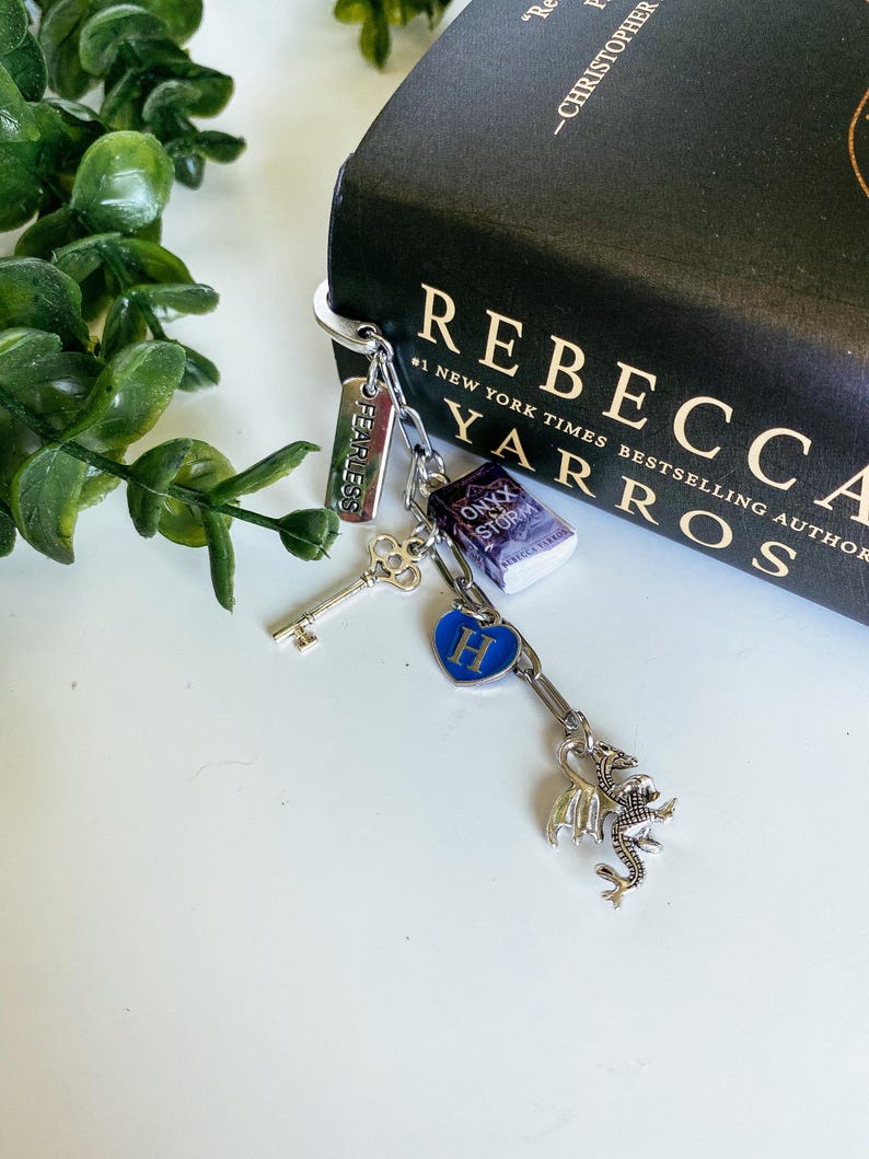 May include: A silver-toned metal keychain featuring multiple charms: a rectangular tag engraved with the word FEARLESS, a miniature book titled Onyx Storm, a silver skeleton key, a blue heart charm with the letter H, and a detailed dragon figure.