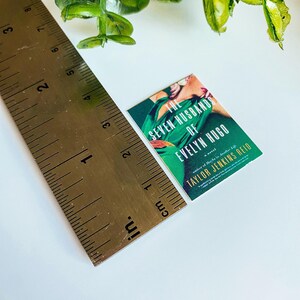 Puede incluir: Un libro de bolsillo titulado "The Seven Husbands of Evelyn Hugo" de Taylor Jenkins Reid. La portada presenta a una mujer con un vestido verde en una pose dram&aacute;tica.