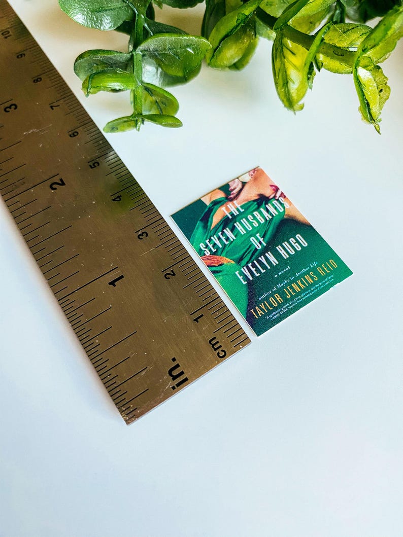 Puede incluir: Un libro de bolsillo titulado "The Seven Husbands of Evelyn Hugo" de Taylor Jenkins Reid. La portada presenta a una mujer con un vestido verde en una pose dram&aacute;tica.