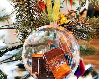 Puede incluir: Un adorno esférico de plástico transparente que cuelga de una cinta verde, relleno de réplicas de libros en miniatura. Las portadas visibles incluyen títulos como Caught Up, The Do-Over y un artículo de Tom Ford, con varios diseños gráficos coloridos.
