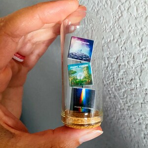 May include: A clear glass vial with a cork base, containing three miniature album covers. The album covers feature colorful artwork and the band name "Imagine Dragons". The vial is held in a hand.