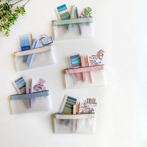 May include: Four clear zippered pouches with different colored trim, each filled with colorful stationery, including pens, highlighters, and sticky notes. The pouches are on a white surface.