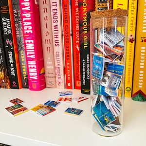 May include: A glass jar filled with miniature books, some with colorful covers and others with white covers. The jar is sitting on a white surface in front of a bookshelf with a variety of books.