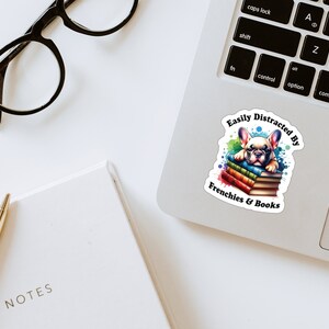 May include: A sticker featuring a cartoon French bulldog puppy lying on a stack of colourful books. The sticker has the text "Easily Distracted By Frenchies & Books" around the puppy.