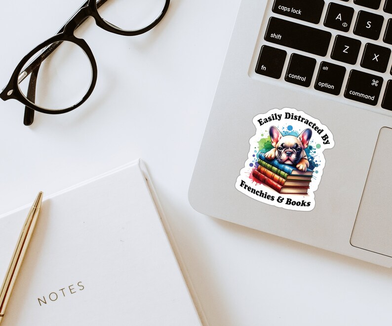May include: A sticker featuring a cartoon French bulldog puppy lying on a stack of colourful books. The sticker has the text "Easily Distracted By Frenchies & Books" around the puppy.