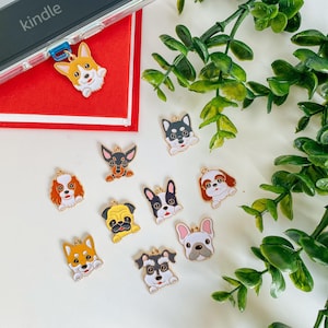 May include: A collection of ten gold-colored metal charms featuring different dog breeds. The charms are on a white surface with green foliage in the background.
