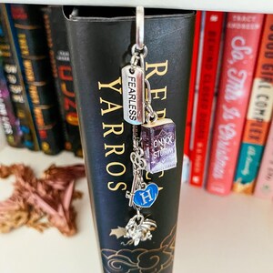 May include: A silver-toned metal bookmark charm hanging from the top of a black book spine. The charm features a rectangular tag engraved with the word FEARLESS, a miniature book titled ONYX STORM, a skeleton key, a blue heart with the letter H, and a dragon.