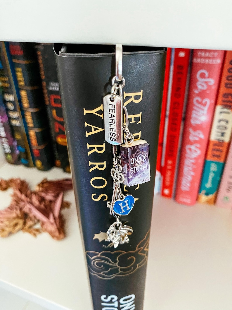 May include: A silver-toned metal bookmark charm hanging from the top of a black book spine. The charm features a rectangular tag engraved with the word FEARLESS, a miniature book titled ONYX STORM, a skeleton key, a blue heart with the letter H, and a dragon.