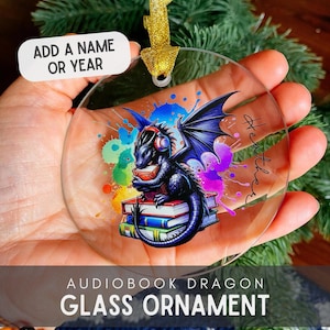 May include: A clear glass bauble featuring a black dragon wearing headphones and holding a book. The dragon is sitting on a stack of books with a colourful watercolour background. The bauble has a gold ribbon attached to the top. The text "Add a name or year" is visible on a white label.