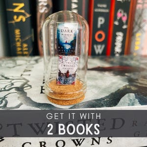 May include: A glass cloche with a cork base, containing two miniature paperback books. The books are titled "On a Dark Window" and "Two Twisted Crowns". The text "GET IT WITH 2 BOOKS" is printed below the cloche.
