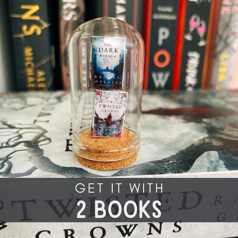 May include: A glass cloche with a cork base, containing two miniature paperback books. The books are titled "On a Dark Window" and "Two Twisted Crowns". The text "GET IT WITH 2 BOOKS" is printed below the cloche.