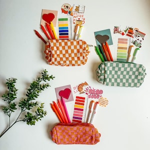 May include: Three different checkered pencil cases with colourful pens and pencils inside. Each case has a heart-shaped sticker on the front. The text on the stickers reads "Read More Books" and "Let's Read More Books".