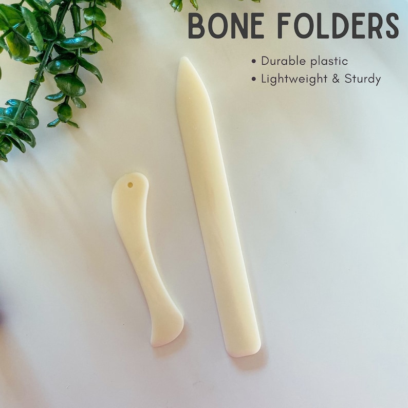 May include: Two off-white bone folders, one long and straight, the other curved with a small hole. The text "BONE FOLDERS" is at the top, with bullet points below stating "Durable plastic" and "Lightweight & Sturdy".