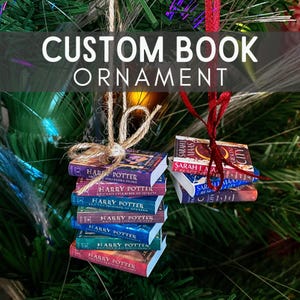 May include: Two miniature book ornaments hanging by strings. One stack features seven Harry Potter books with colourful spines, tied with tan twine. The other stack includes three Sarah J. Maas books with red and blue spines, tied with a red ribbon.