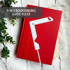 May include: White 5-in-1 bookbinding guide ruler with markings in inches and centimetres. The ruler is on top of a red book.