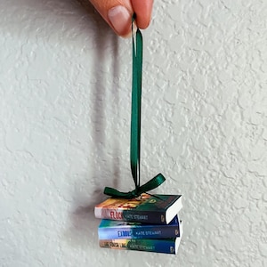 May include: A miniature ornament featuring a stack of three books tied with a dark green satin ribbon. The book covers display the titles Flock, Exodus, and The Finish Line by Kate Stewart. The stack is suspended by a matching green ribbon loop.