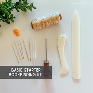 May include: A basic starter bookbinding kit with needles, thread, awl, bone folders, and other tools. The kit includes a spool of beige thread, several needles, and two white bone folders. The text "BASIC STARTER BOOKBINDING KIT" is displayed.