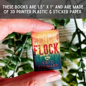 May include: A miniature book measuring 3.81 cm by 2.54 cm, crafted from 3D printer plastic and sticker paper. The cover features the title Flock by Kate Stewart, with a sunset sky over a forest landscape and the text Can you keep a secret?