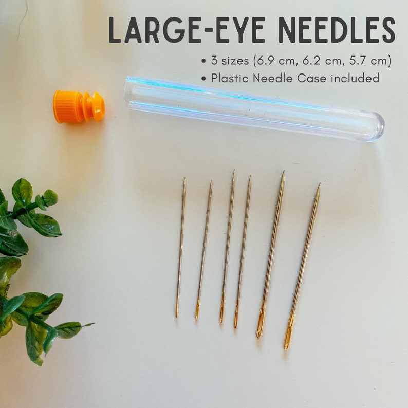 May include: A set of large-eye needles with a clear plastic case and an orange cap. The text reads "LARGE-EYE NEEDLES" and lists three sizes: 6.9 cm, 6.2 cm, and 5.7 cm. The needles are gold-toned.