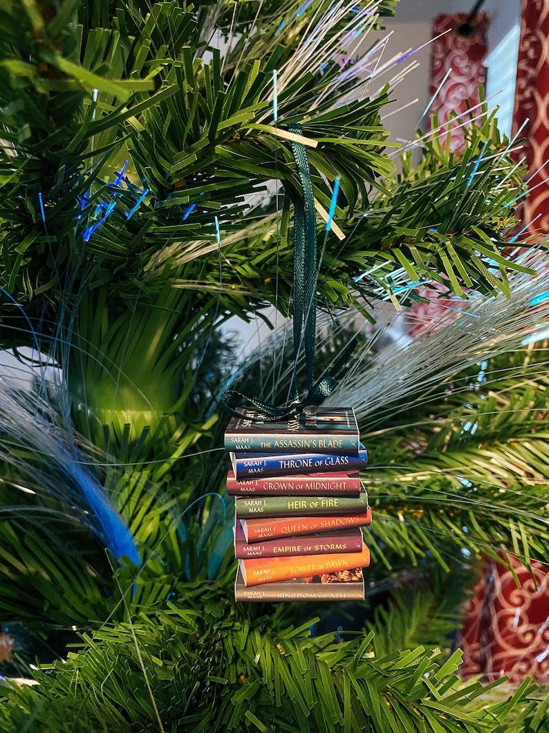 May include: A miniature stack of eight book ornaments by Sarah J. Maas, including The Assassin's Blade, Throne of Glass, Crown of Midnight, Heir of Fire, Queen of Shadows, Empire of Storms, Tower of Dawn, and Kingdom of Ash, hanging by a green ribbon.