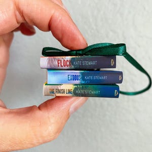 May include: A miniature stack of three books titled Flock, Exodus, and The Finish Line by Kate Stewart. The books are bound together with a dark green ribbon loop at the top, featuring colourful spines with white text and a small gold logo.