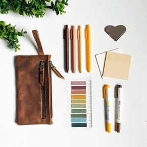 Puede incluir: Un estuche de lápices de cuero marrón con tres compartimentos con cremallera. El estuche está abierto y contiene cinco bolígrafos, cuatro de los cuales son de color marrón, rojo y amarillo. También hay una tabla de colores con una variedad de colores y un par de rotuladores.