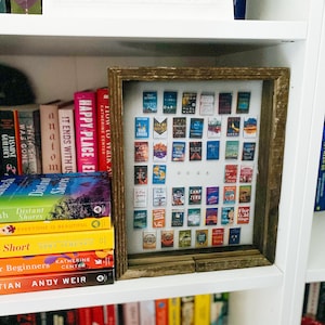 May include: A white bookshelf with a framed display of small book covers. The book covers are arranged in a grid pattern and feature a variety of titles. The bookshelf is also filled with other books, including some with colourful spines.