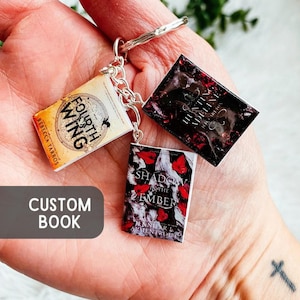 May include: A keychain featuring three miniature book charms. The books are titled "Fourth Wing", "Hunting Adeline", and "Shadow in the Ember". The keychain also has a tag that says "Custom Book".