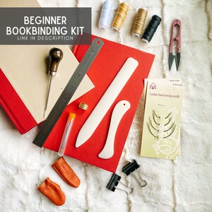 May include: Beginner bookbinding kit with tools and supplies including a bone folder, awl, thimble, needles, thread, and leather finger cots.  The kit is laid out on a white faux fur surface.  The text on the image reads "Beginner Bookbinding Kit Link in Description."