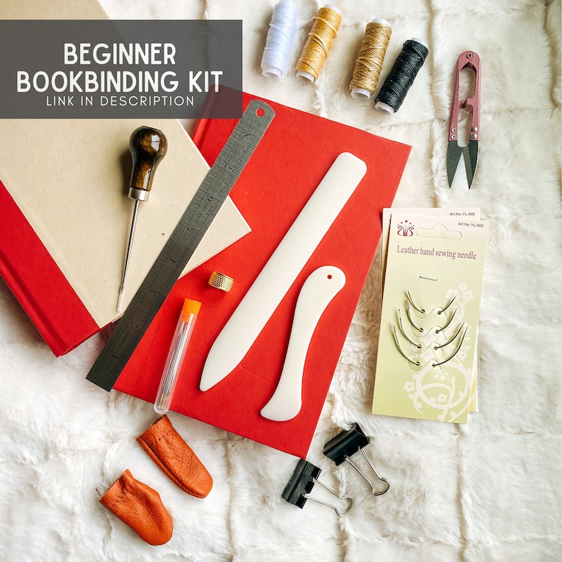 May include: Beginner bookbinding kit with tools and supplies including a bone folder, awl, thimble, needles, thread, and leather finger cots.  The kit is laid out on a white faux fur surface.  The text on the image reads "Beginner Bookbinding Kit Link in Description."
