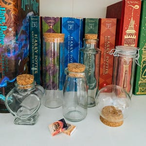May include: Collection of clear glass bottles and jars with cork stoppers, displayed in front of a row of Harry Potter books. The bottles vary in shape, including a heart-shaped bottle and a spherical jar. Two small books are in front of the bottles.
