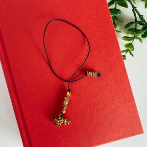 May include: A black bookmark with gold beads and a lion charm. The bookmark is on a red book.