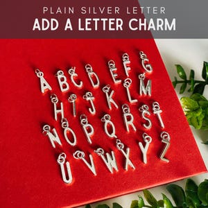 May include: A collection of silver-toned metal letter charms featuring the alphabet from A to Z. Each individual charm includes a small circular jump ring at the top for attachment. The text PLAIN SILVER LETTER ADD A LETTER CHARM is visible.