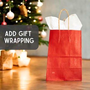 May include: A vertical red paper gift bag with a ribbed texture and brown twisted paper handles. White tissue paper protrudes from the top opening. A dark gray rectangular overlay features the white text ADD GIFT WRAPPING.