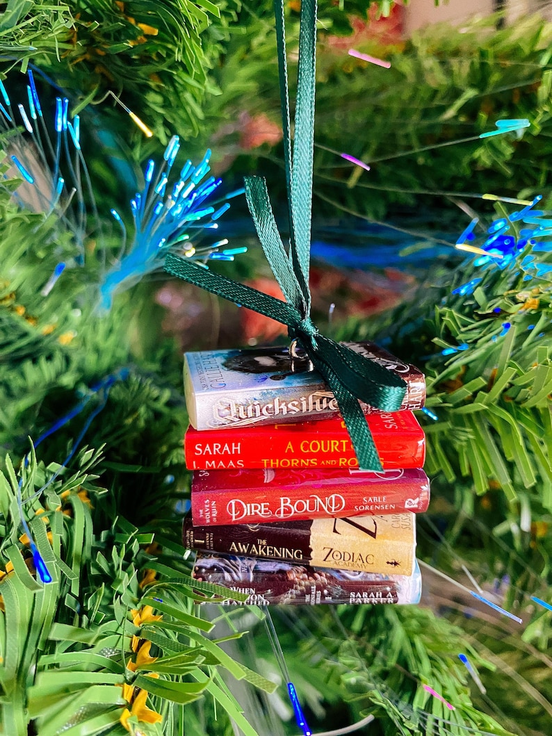 May include: A miniature stack of five book ornaments tied together with a dark green satin ribbon. The spines feature titles including Quicksilver, A Court of Thorns and Roses, Dire Bound, The Awakening Zodiac Academy, and When Hatched.
