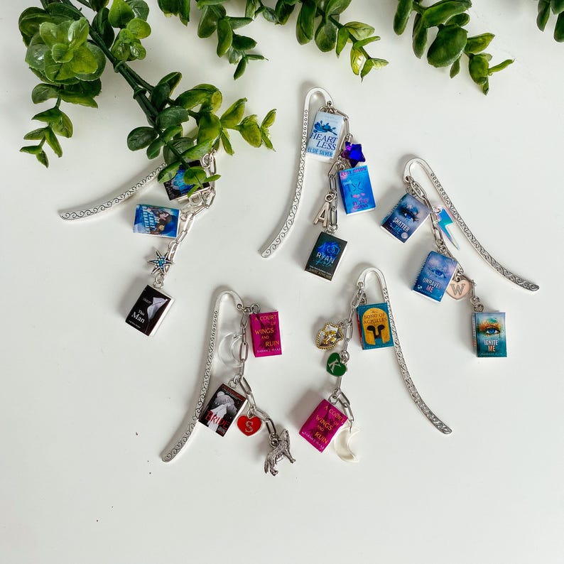 May include: Five silver metal bookmarks, each featuring a long, flat, patterned stem and a chain attachment. The chains hold miniature book charms with various titles and covers, along with small decorative charms like stars, hearts, and letters.