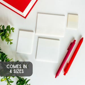 May include: White sticky notes in four sizes are arranged on a white surface, accompanied by two pens, one red and one magenta. A red book and greenery are in the background. The text "COMES IN 4 SIZES" is visible.
