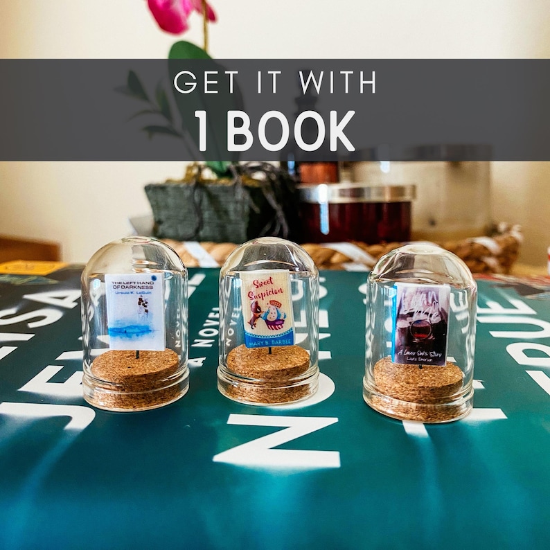May include: Three glass cloche domes with cork bases, each containing a miniature book. The books are titled "The Light We Carry", "Sweet Suspicion", and "A Love Story".