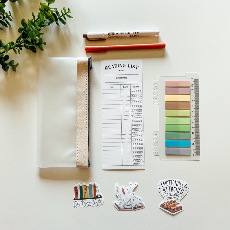 May include: A clear plastic pencil pouch with a beige zipper pull, a reading list printable with a title, date, and rating section, a set of colorful sticky notes, and three book-themed stickers. The stickers feature a stack of books with the text "One More Chapter", a book with flowers, and a book with the text "Emotionally Attached to Fictional Characters".