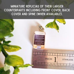 May include: A miniature book charm featuring a brown back cover with text and a barcode, attached to a silver jump ring. A stainless steel ruler is positioned underneath, showing the charm measures approximately 0.75 inches in height.