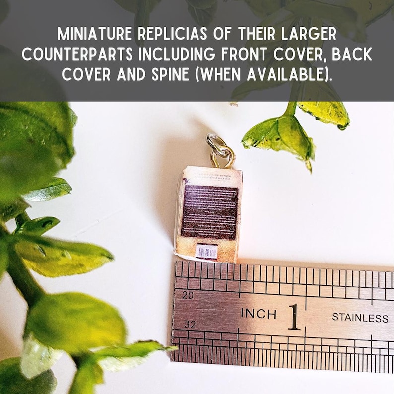 May include: A miniature book charm featuring a brown back cover with text and a barcode, attached to a silver jump ring. A stainless steel ruler is positioned underneath, showing the charm measures approximately 0.75 inches in height.
