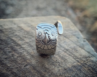 Puede incluir: Un anillo de plata con un diseño de búho. Los ojos del búho son grandes y redondos, y su pico es pequeño y puntiagudo. El anillo está hecho de una sola pieza de metal, y las alas del búho están enroscadas alrededor de la banda del anillo.