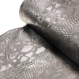 May include: Close-up of a silver snakeskin fabric with a textured, embossed pattern. The fabric is folded in half, showing the texture from both sides.