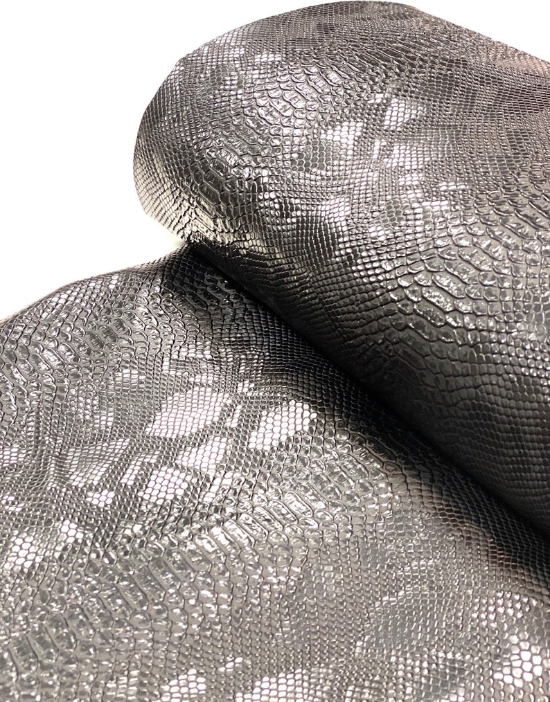May include: Close-up of a silver snakeskin fabric with a textured, embossed pattern. The fabric is folded in half, showing the texture from both sides.