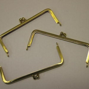 May include: Three gold-colored purse frames with a rectangular shape and a kiss-lock clasp. The frames have a shiny finish and are designed for crafting handbags or clutches. The metal frames are shown against a neutral background.