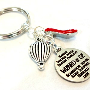 May include: Silver keychain with a red ruby slipper charm, a hot air balloon charm, and a round charm with the words "Wizard of Oz" and a list of characters and items from the movie.