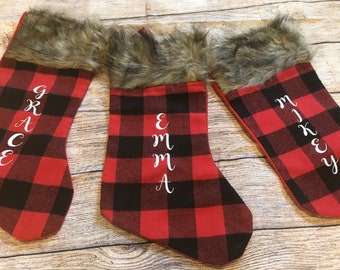 May include: Three red and black buffalo plaid Christmas stockings with faux fur trim. The stockings are personalized with the names "Grace", "Emma", and "Mikey".