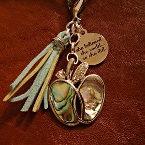 May include: Silver keychain with a charm of an abalone shell apple with rhinestones, a silver tassel with blue and yellow leather strands, and a round silver tag with the text "she believed she could... so she did".