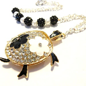 May include: A gold-tone necklace with a black and white turtle pendant. The turtle is covered in clear rhinestones and has a white flower on its shell. The necklace is made of a silver chain with black and white beads.