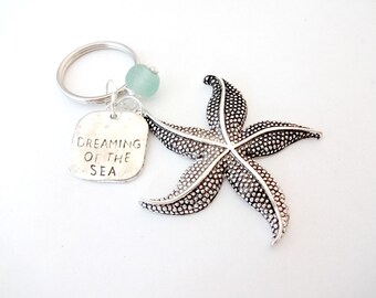 May include: Silver starfish keychain with a charm that reads "Dreaming of the Sea". The charm is attached to a silver key ring with a light blue glass bead.